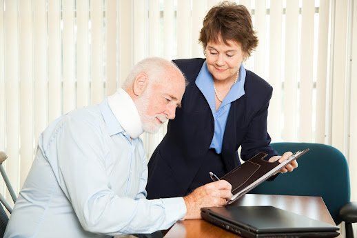 Man with neck brace signing document at desk, assisted by woman in blazer.