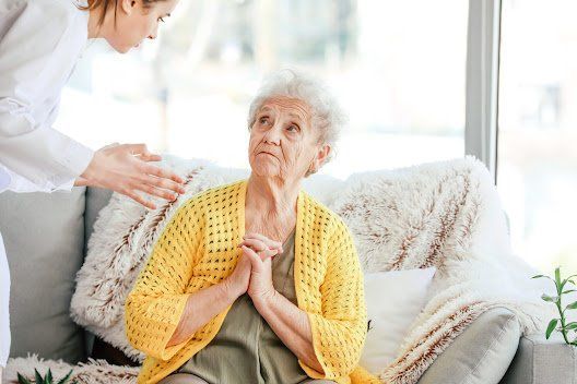 Elderly woman with clasped hands looking upwards, comfort by caregiver on a sofa.