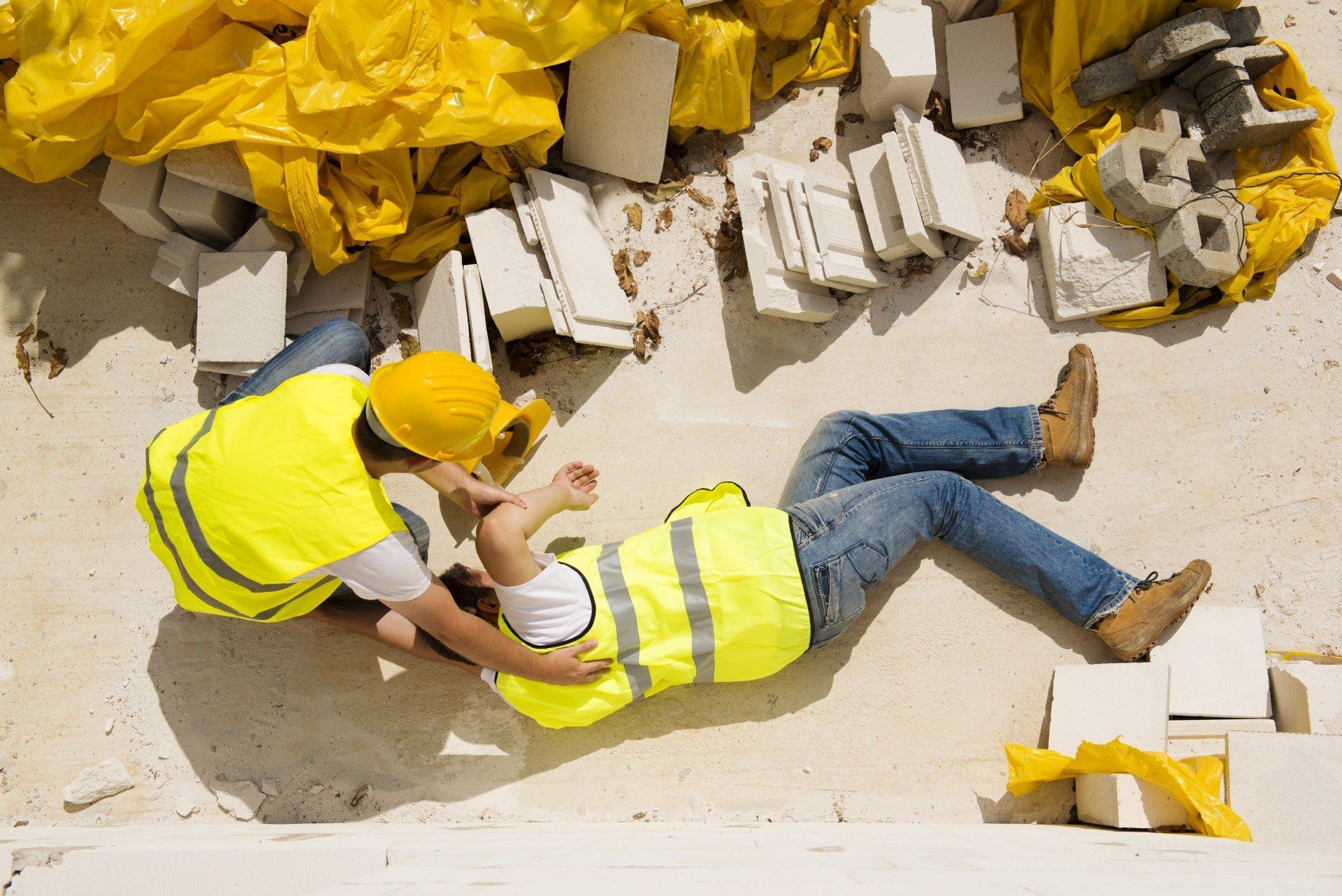 Two construction workers, one helping the other who is on the ground. Yellow vests and hard hats are visible. Debris surrounds them.