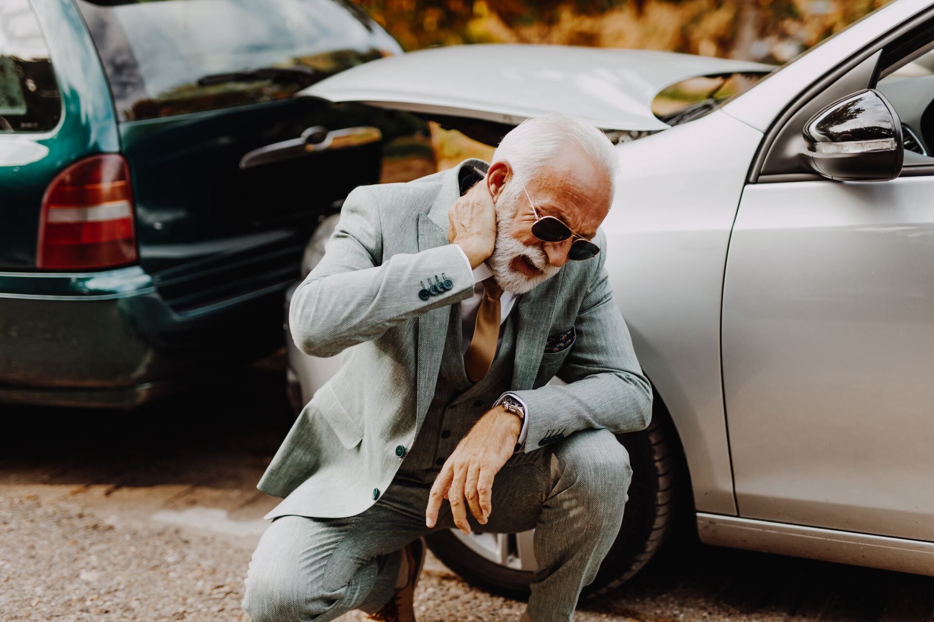 Man crouches by a damaged silver car, clutching his neck after a car accident; a green car is behind.