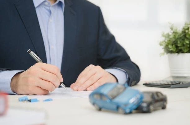 Businessman signing car insurance papers with toy cars, calculator, and plant on office desk.