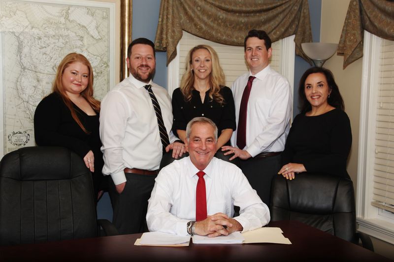 Group of six people in an office. One man sits, others stand behind. All are dressed in business attire.