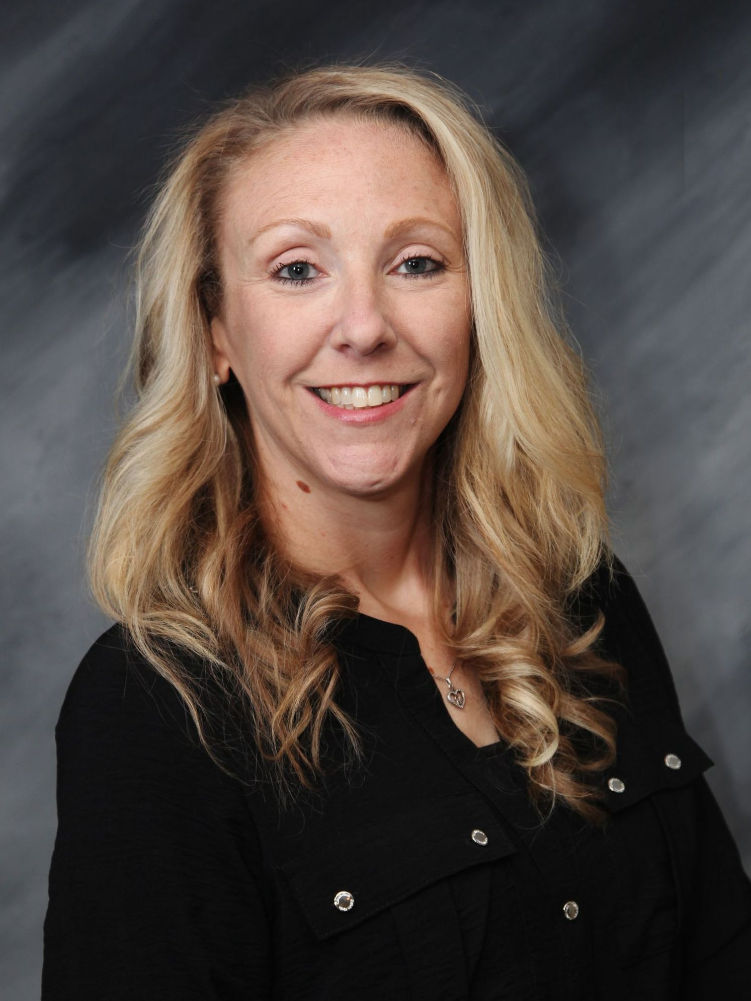 Woman with blonde hair smiling, wearing a black shirt, against a dark gray background.