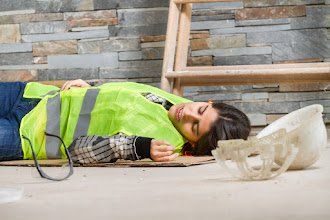 Construction worker lies unconscious on floor near a broken hard hat and a ladder; brick wall background.