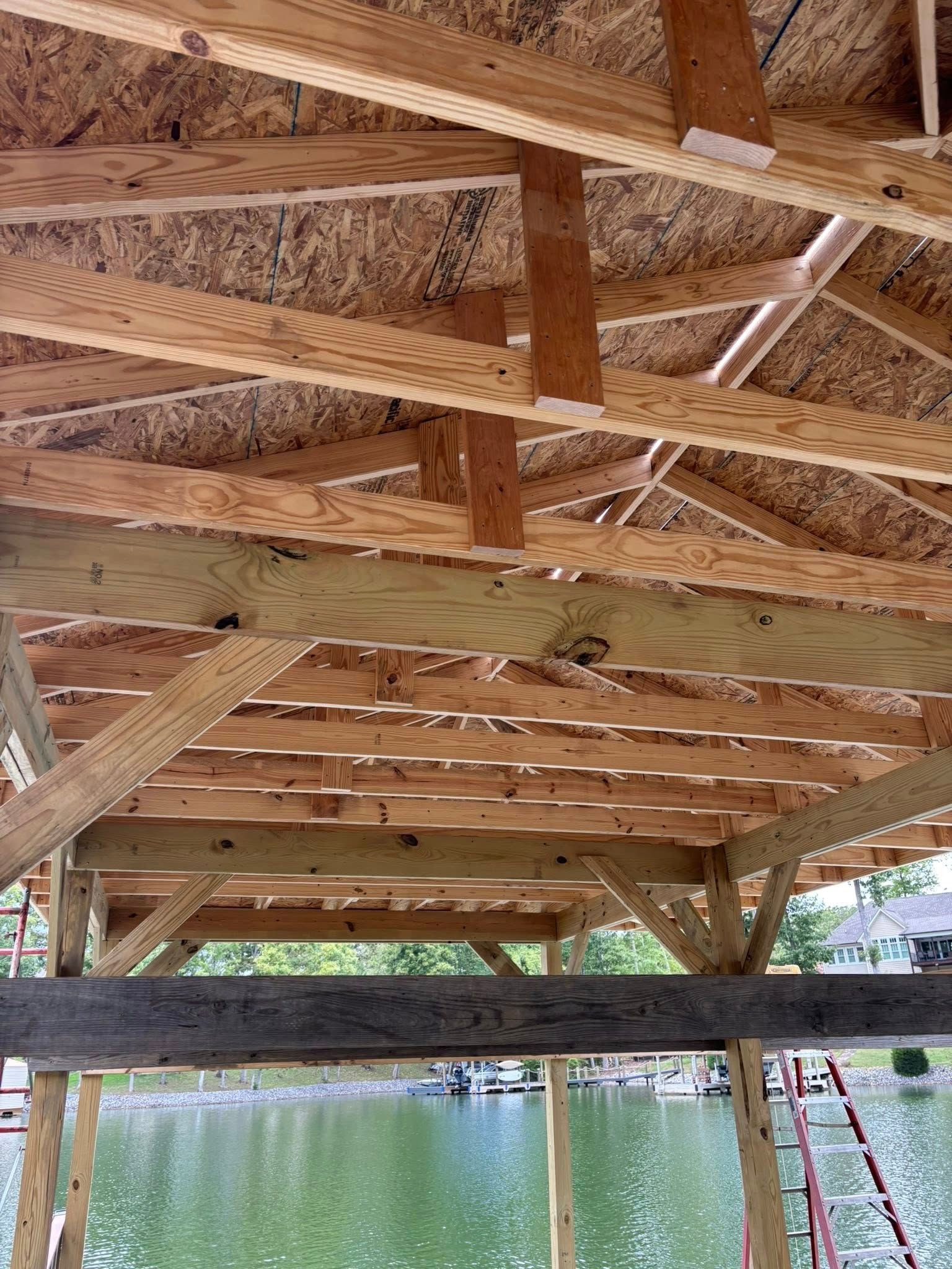 Wooden roof structure of a building under construction, framed with rafters and plywood sheathing, overlooking a lake.