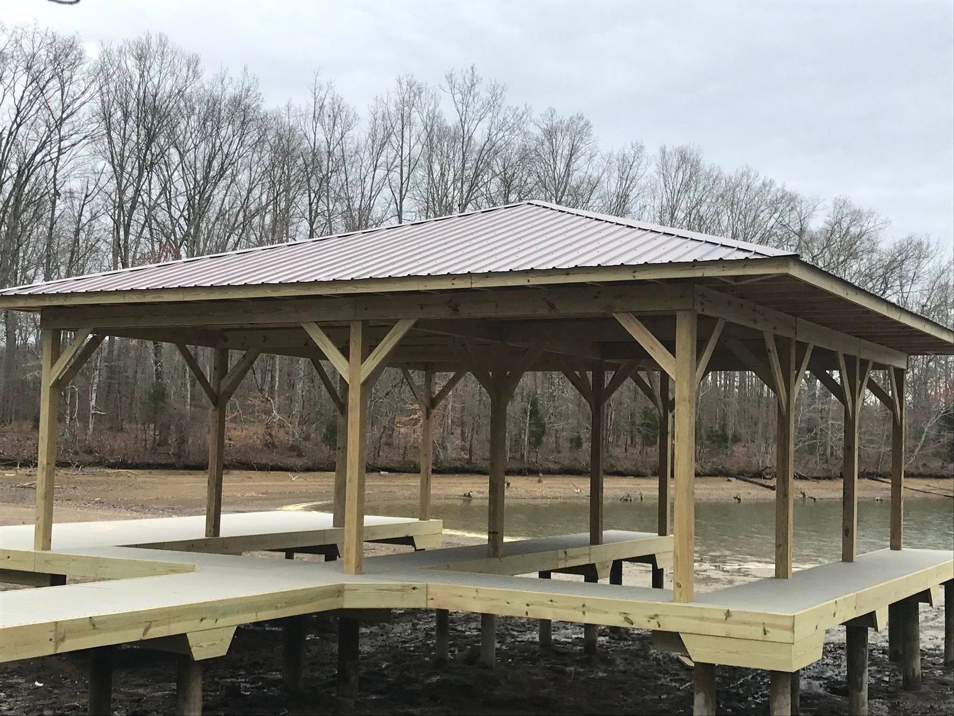 A wooden, covered boat dock on stilts extending over shallow water near a wooded shoreline under an overcast sky.