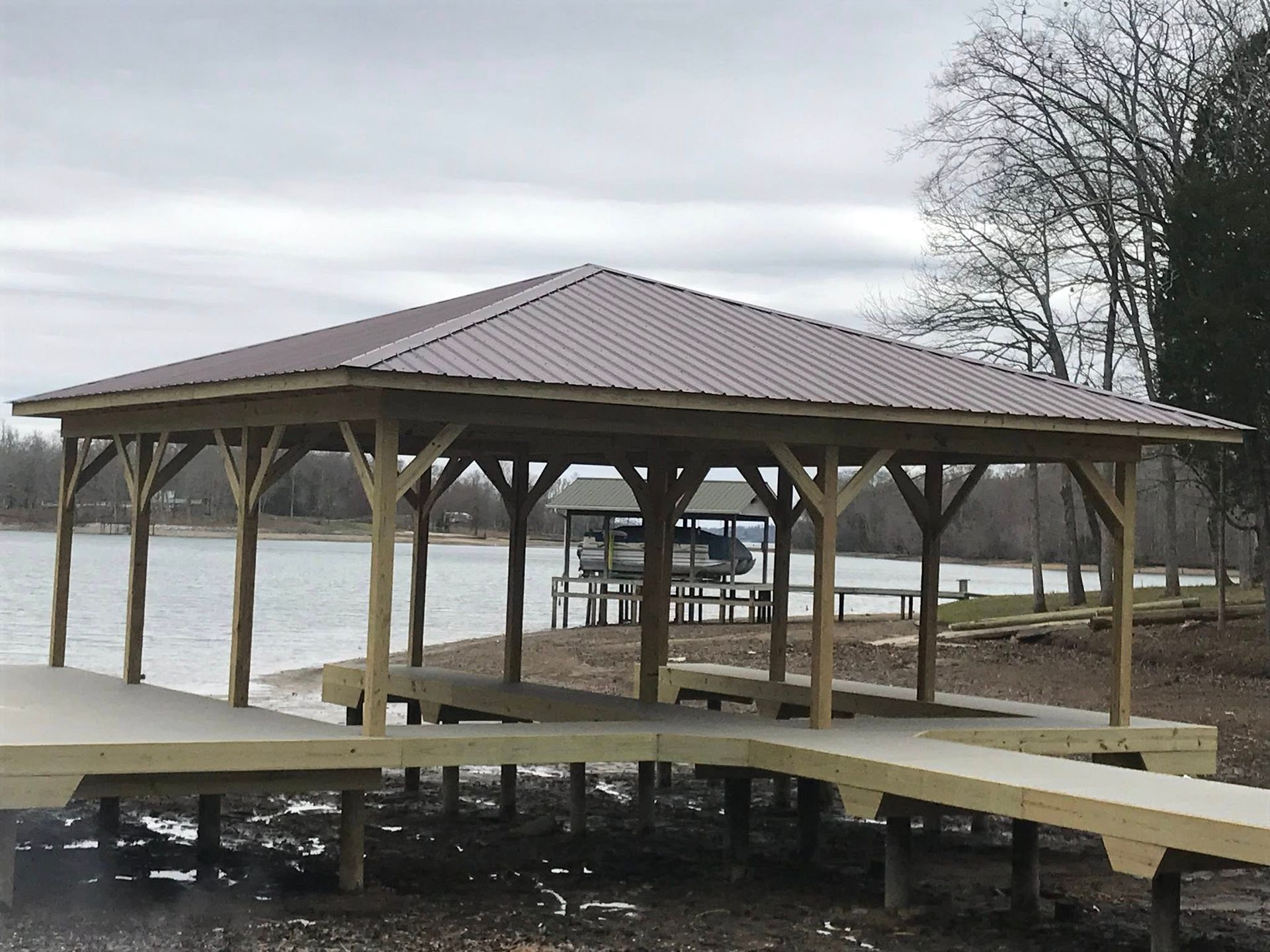 A wooden dock structure with a brown metal roof standing on a muddy shoreline, with a boat lift in the distance.