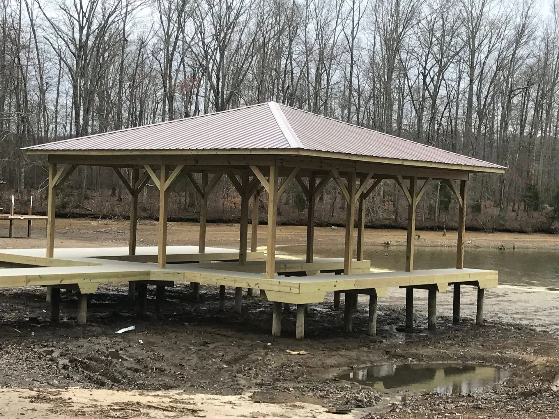 A wooden gazebo with a metal roof stands on pilings over a muddy, dried-up lakebed in front of a forest.