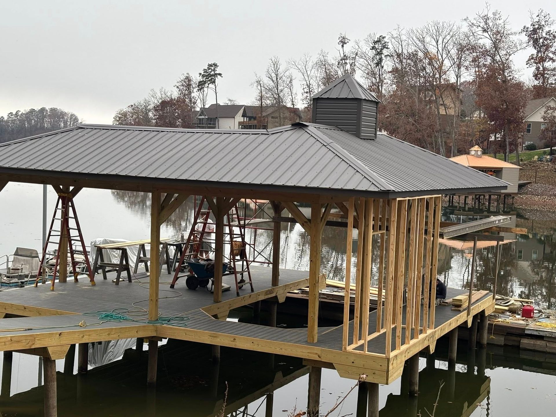 A wooden boat dock structure under construction on a calm lake, featuring a new dark metal roof and an open-framed wall.