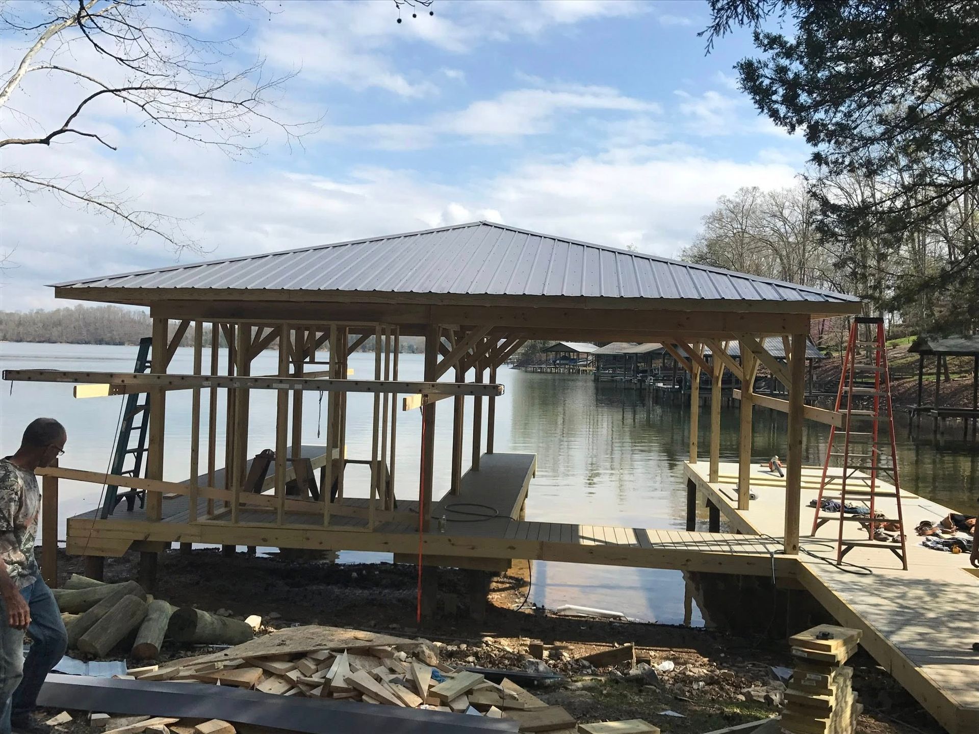 Construction of a wooden boat dock frame with a metal roof on a lake, featuring a person working on the shore nearby.