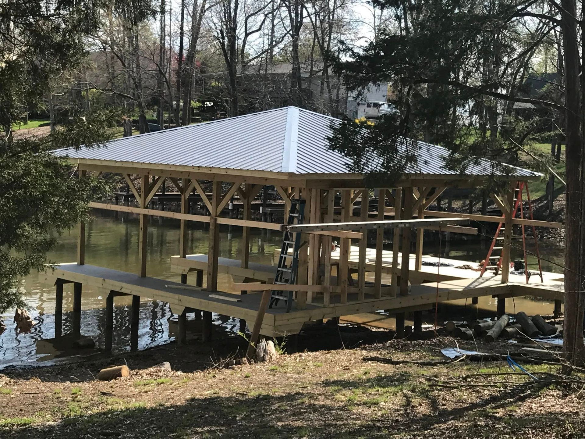 A partially constructed wooden boat dock with a silver metal hip roof, situated on a calm, tree-lined lake.