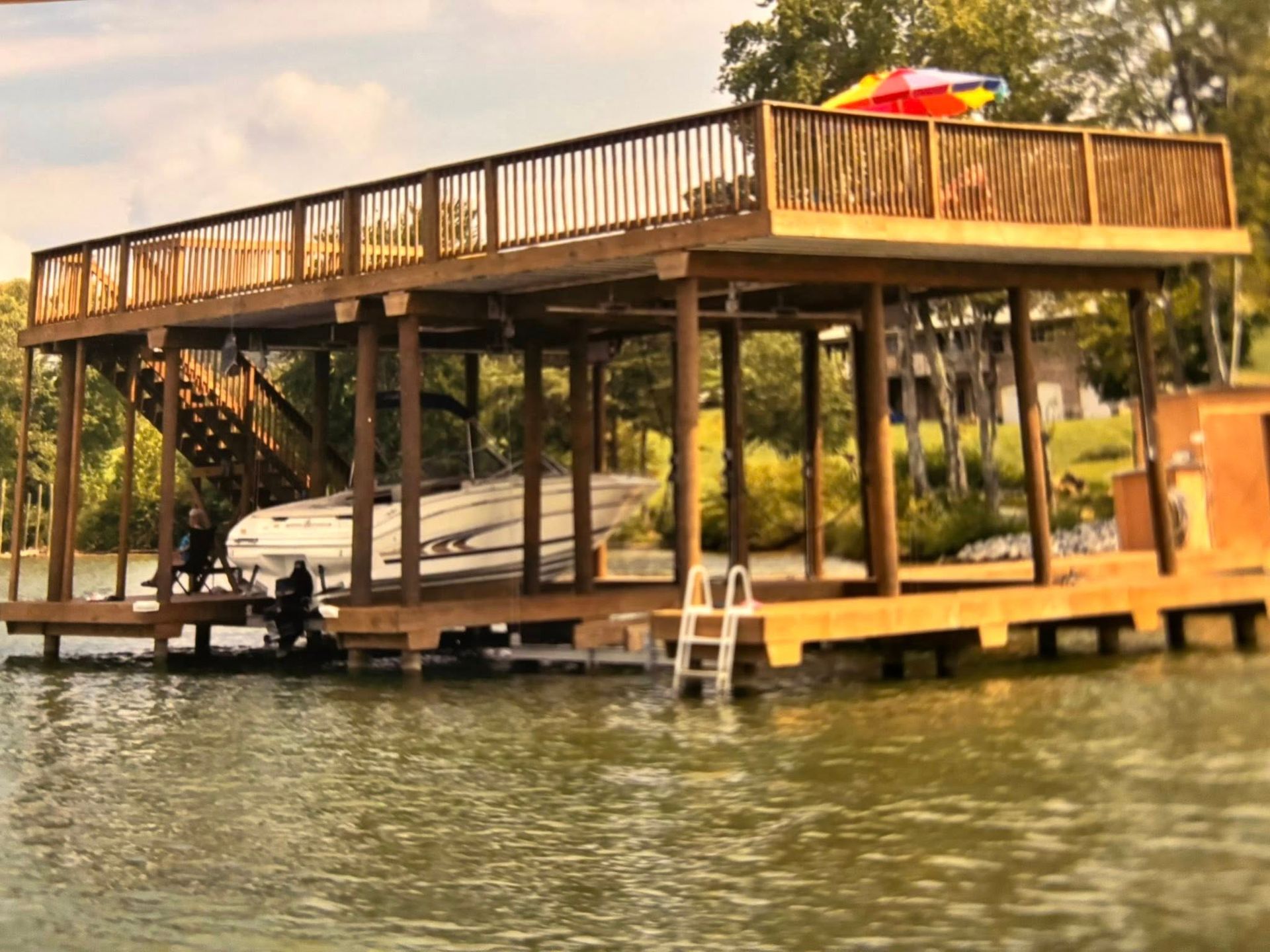 A wooden, two-story boat dock on a lake with a boat moored underneath and a deck above, featuring a small white ladder.