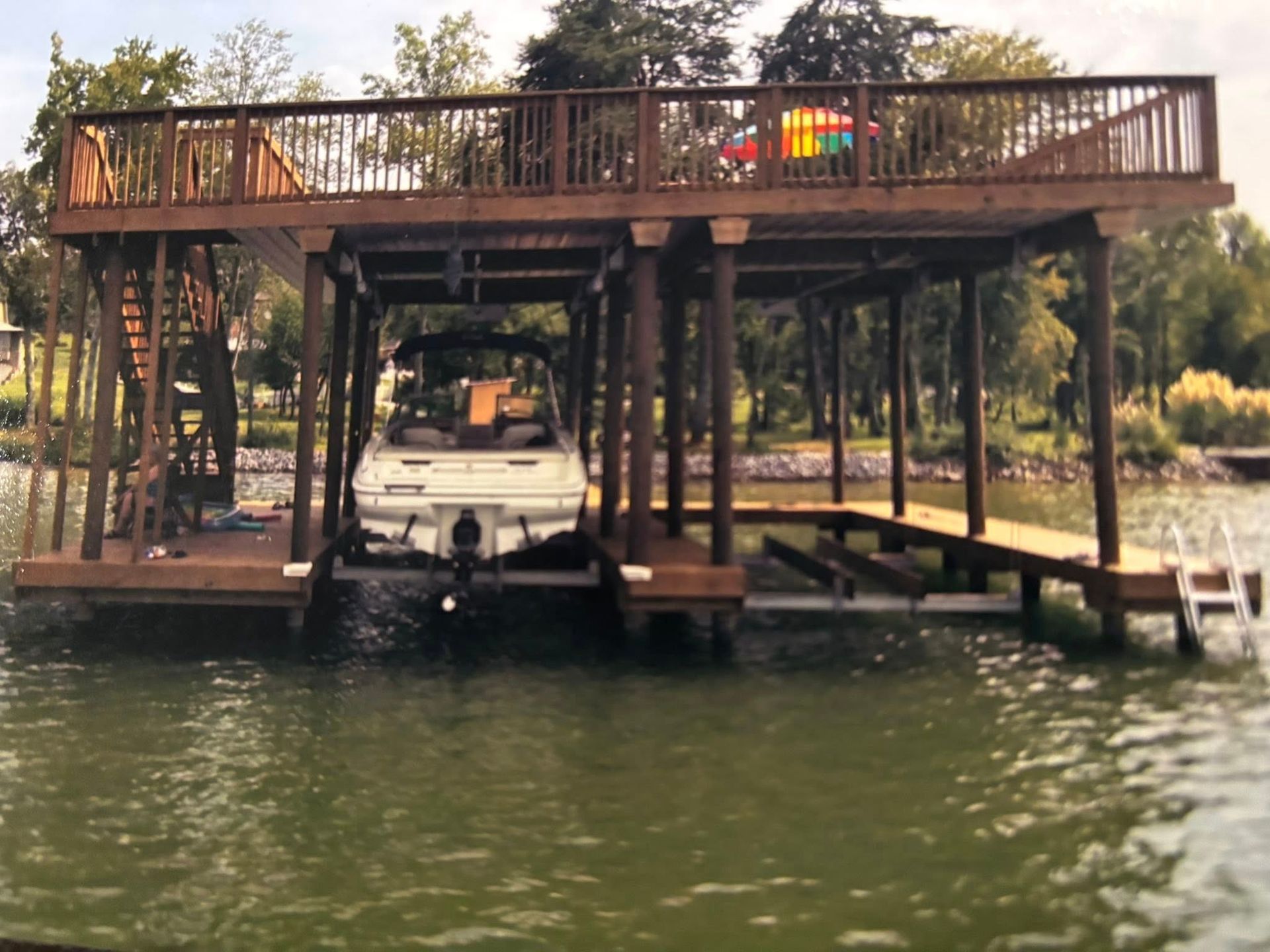 A wooden boat dock on a lake with a boat in one slip, a ladder on the right, and an upper deck with a staircase.