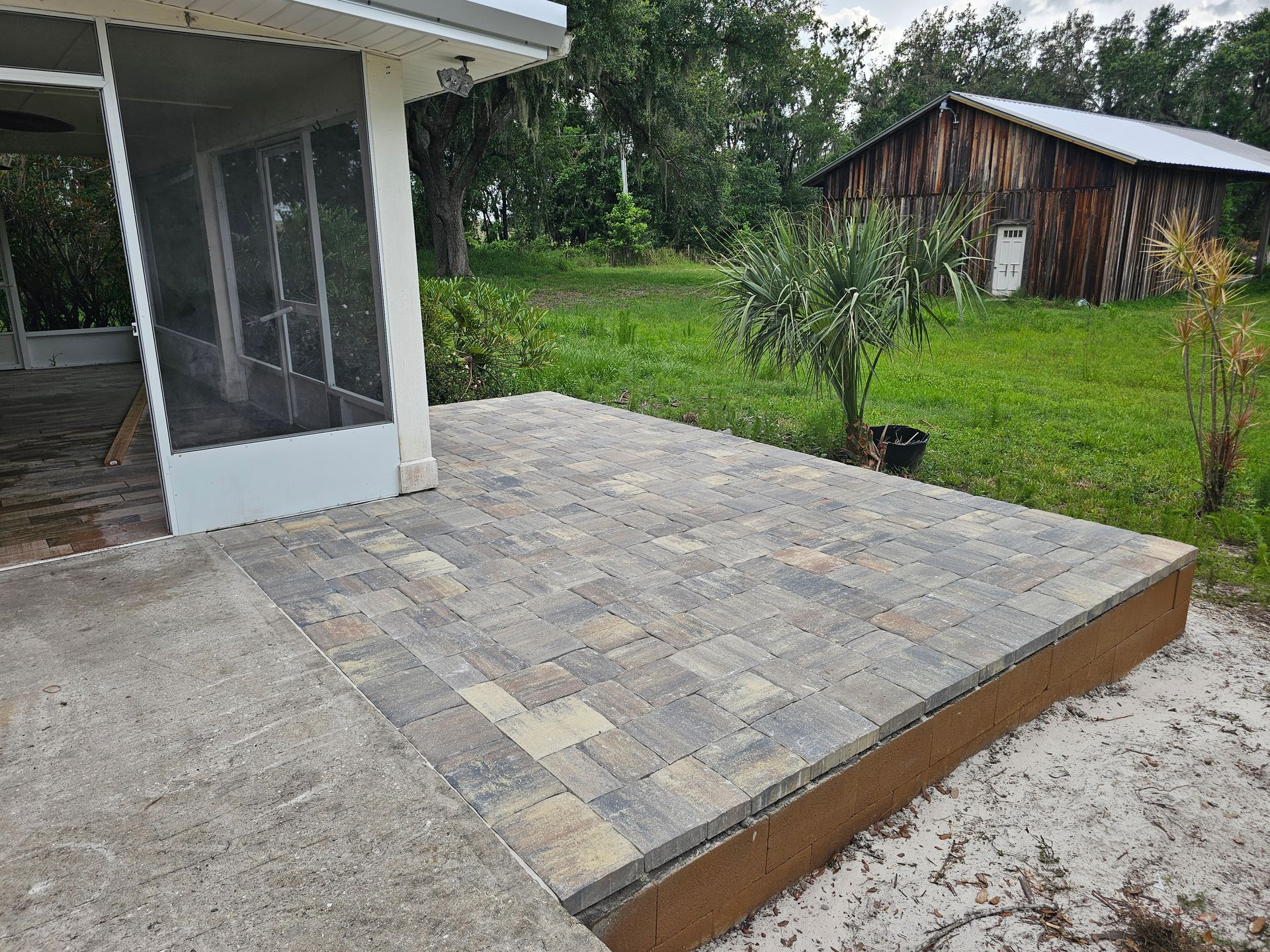 Patio with brick pavers next to a screened porch and grassy yard with a wooden shed.