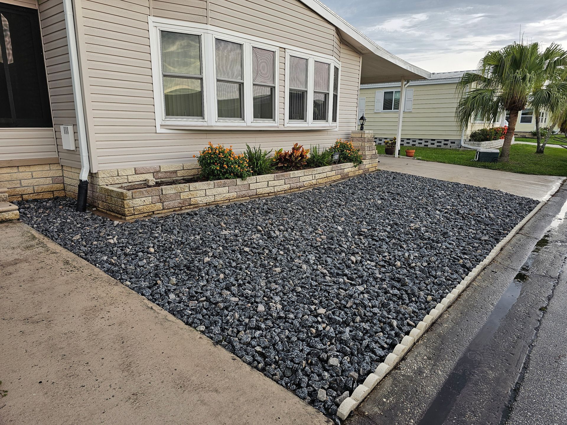 Gravel bed with small plants in front of a house. Concrete sidewalk and street on the right. Overcast sky.