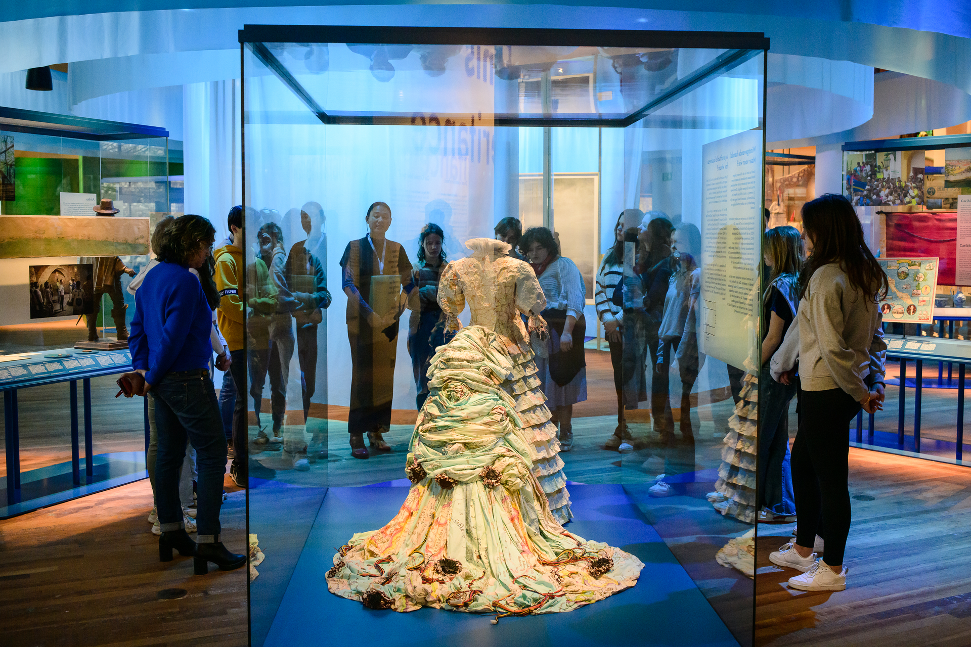 Museumexpositie met een jurk van landkaarten in een glazen vitrine, omringd door bezoekers onder blauwe verlichting.