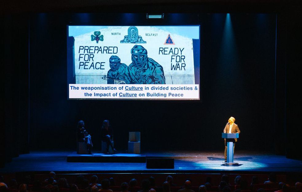 Een persoon spreekt vanaf een podium, op de achtergrond is een groot scherm te zien met een propagandaposter: 