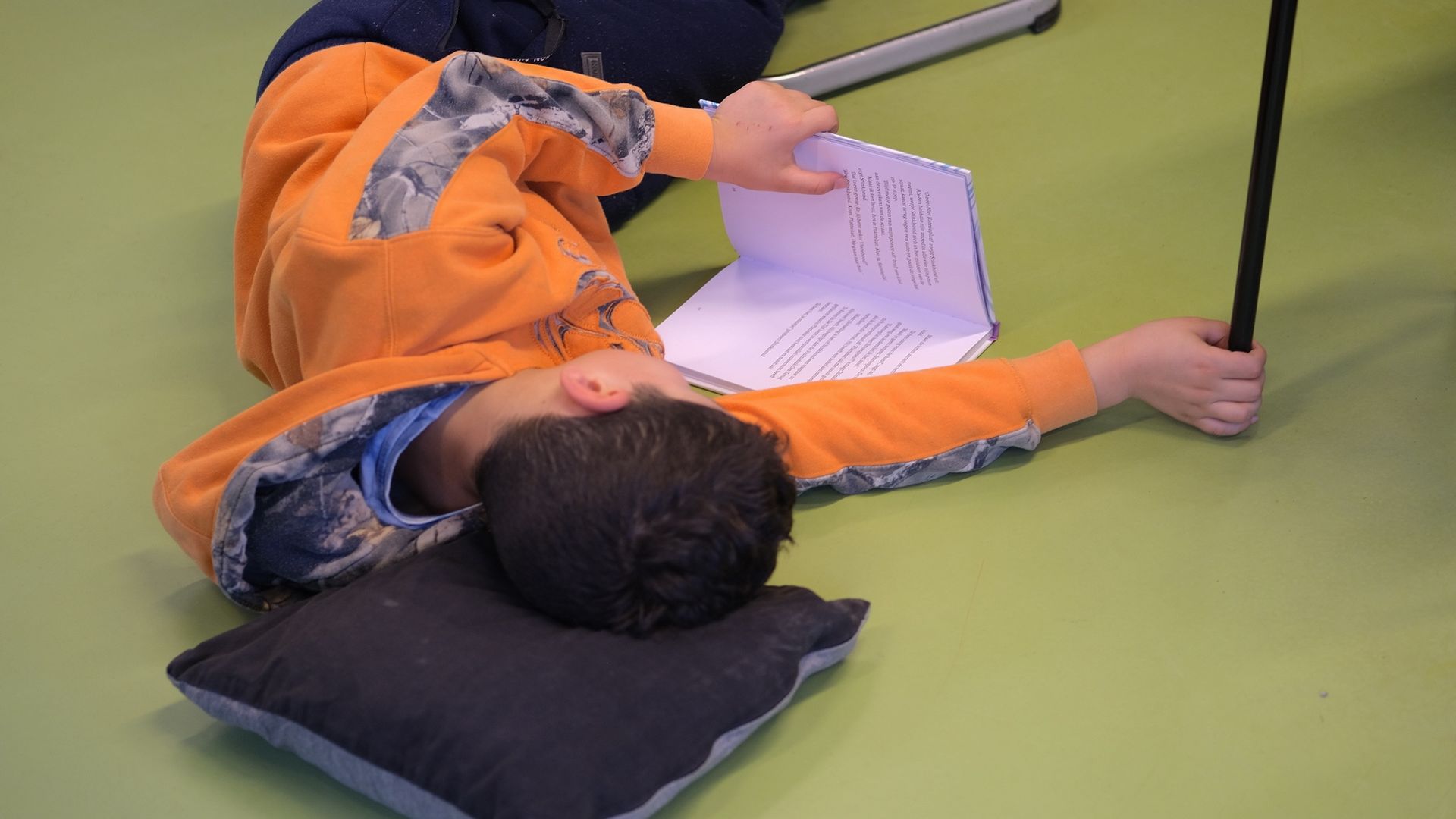 Een persoon ligt op een groene vloer en leest, terwijl hij een zwarte stoelpoot vasthoudt naast een kussen.