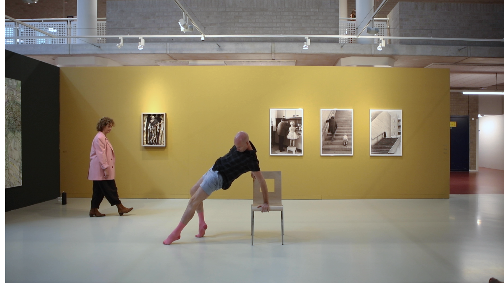 Een man op roze hakken leunt achterover in een stoel in een galerie, een vrouw loopt langs kunstwerken op een gele muur.