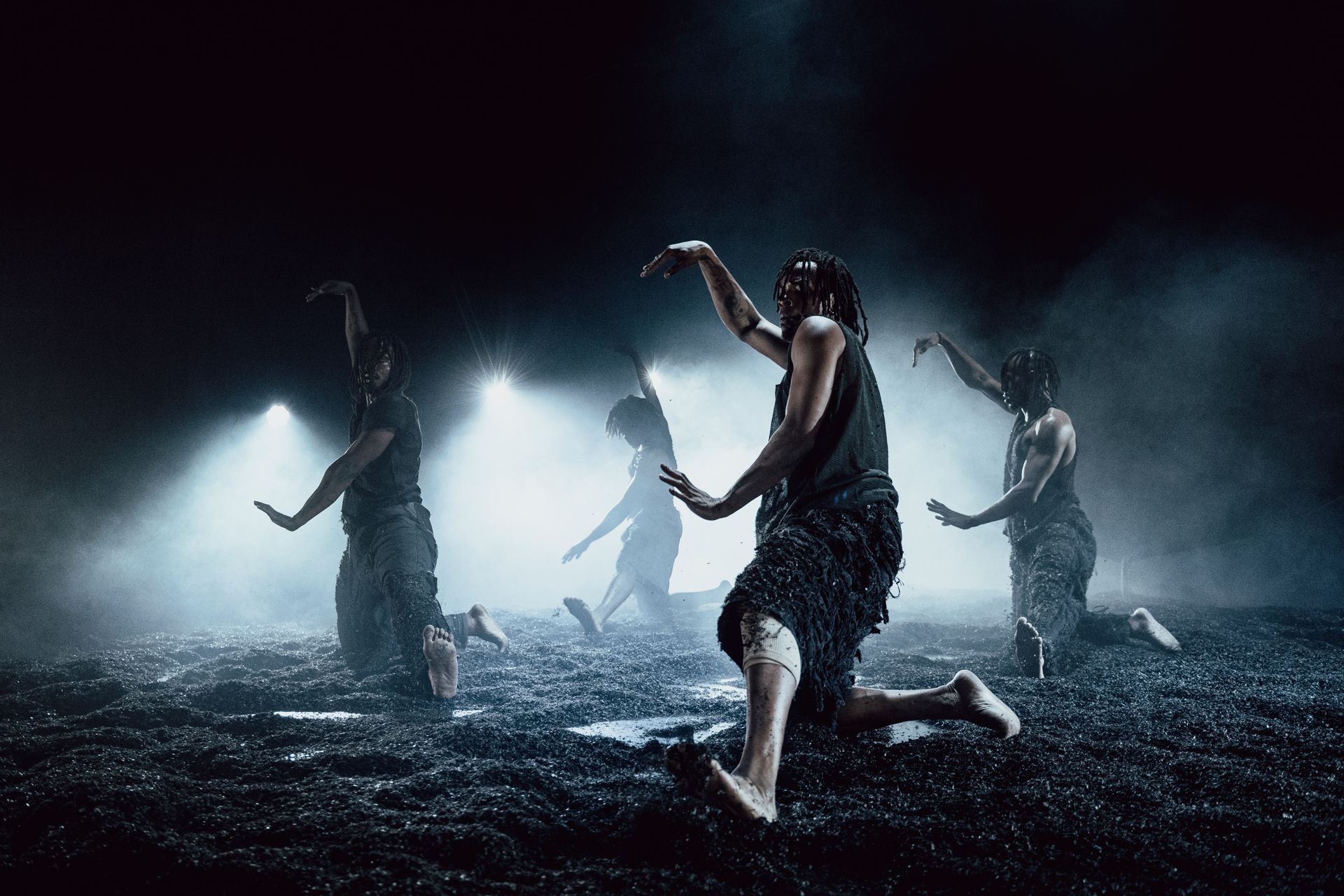 Dansers treden op op een donker podium, onder spotlights. Lichamen in beweging; armen omhoog. Mistige atmosfeer.