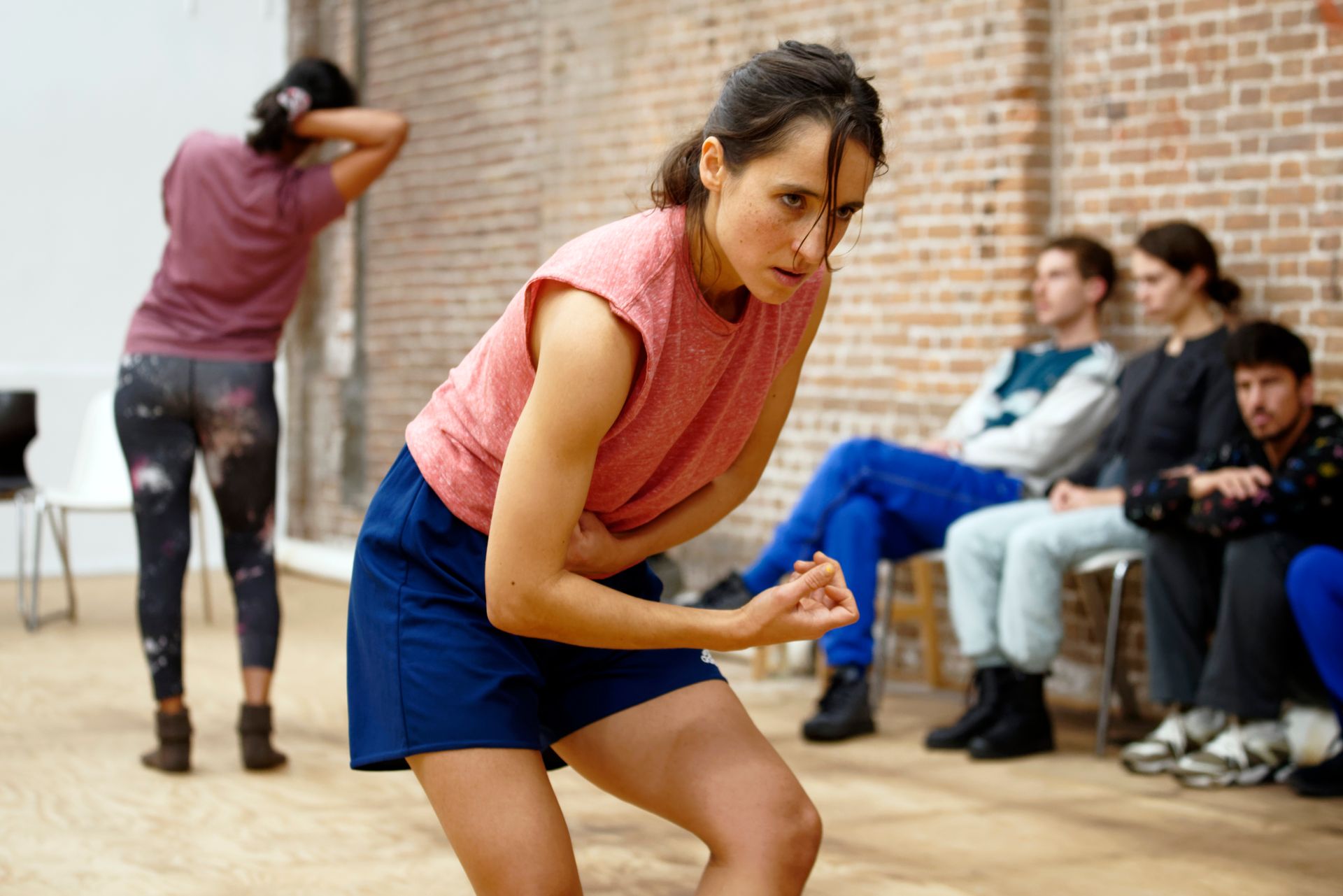 Een vrouw in een perzikkleurig shirt en blauwe korte broek treedt op, anderen kijken toe. 