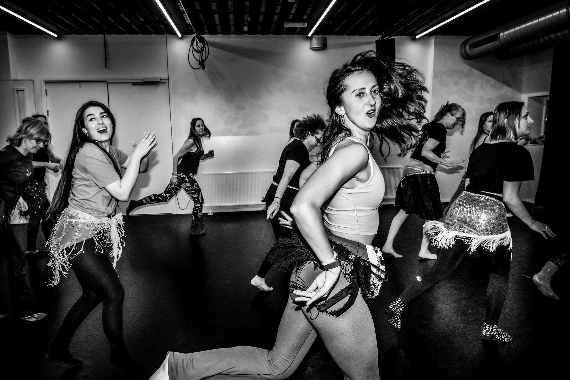Dansers in een studio, tijdens een les. Sommigen dragen heupsjaals en dansen met enthousiaste gezichtsuitdrukkingen.