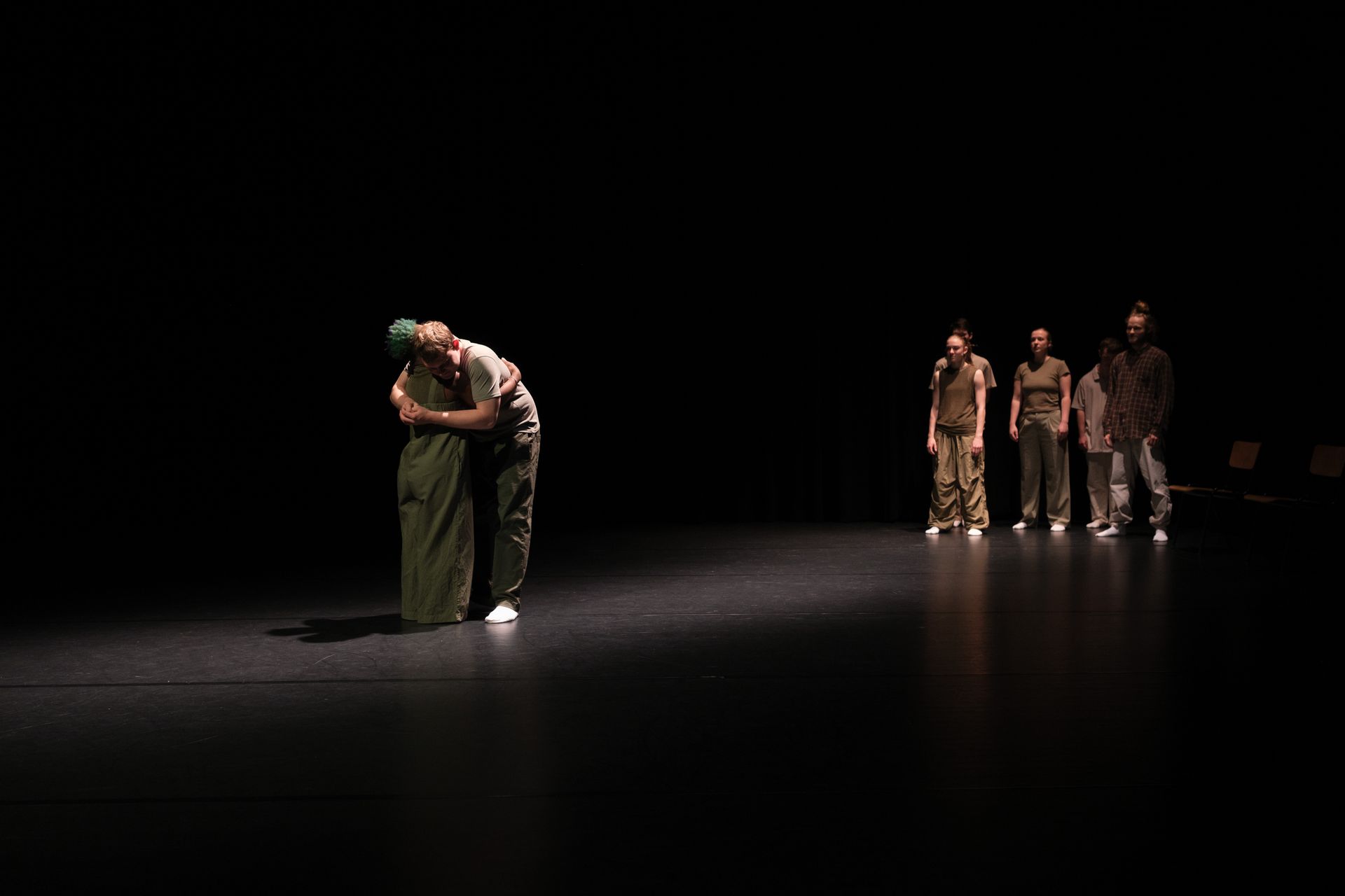 In een schemerig verlicht theater omhelst een persoon een ander; op de achtergrond staan ​​vier figuren.