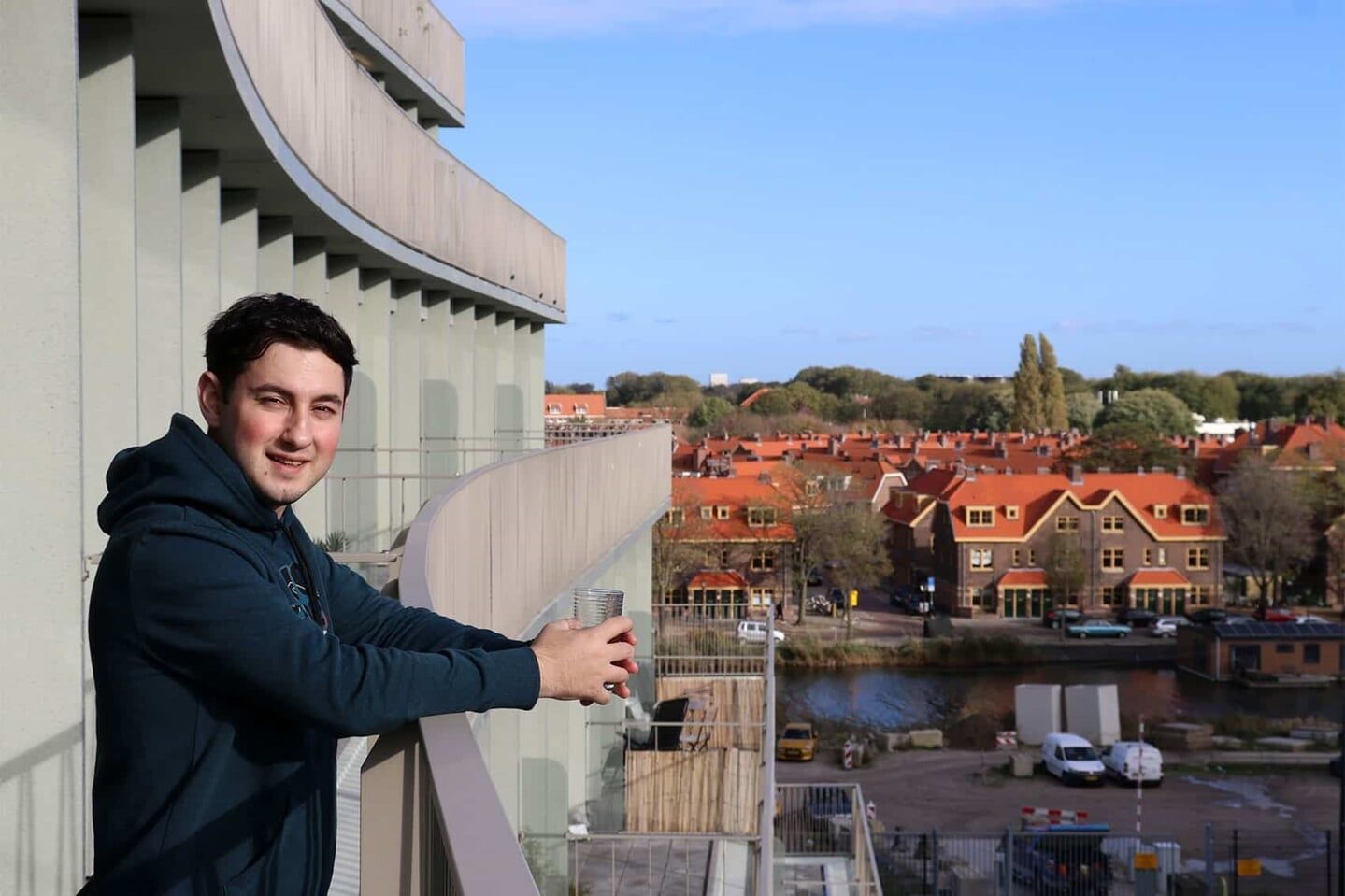 Een man staat op een balkon en kijkt uit over een stadslandschap met gebouwen met rode daken en een blauwe lucht.