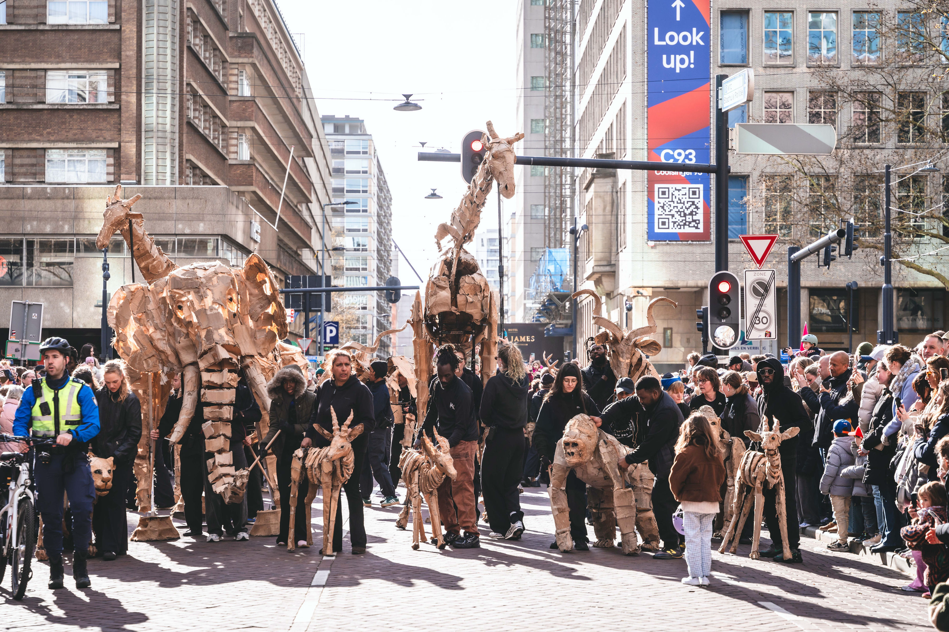 Een parade van papieren giraffen en mensen die door een stadsstraat tussen hoge gebouwen marcheren.