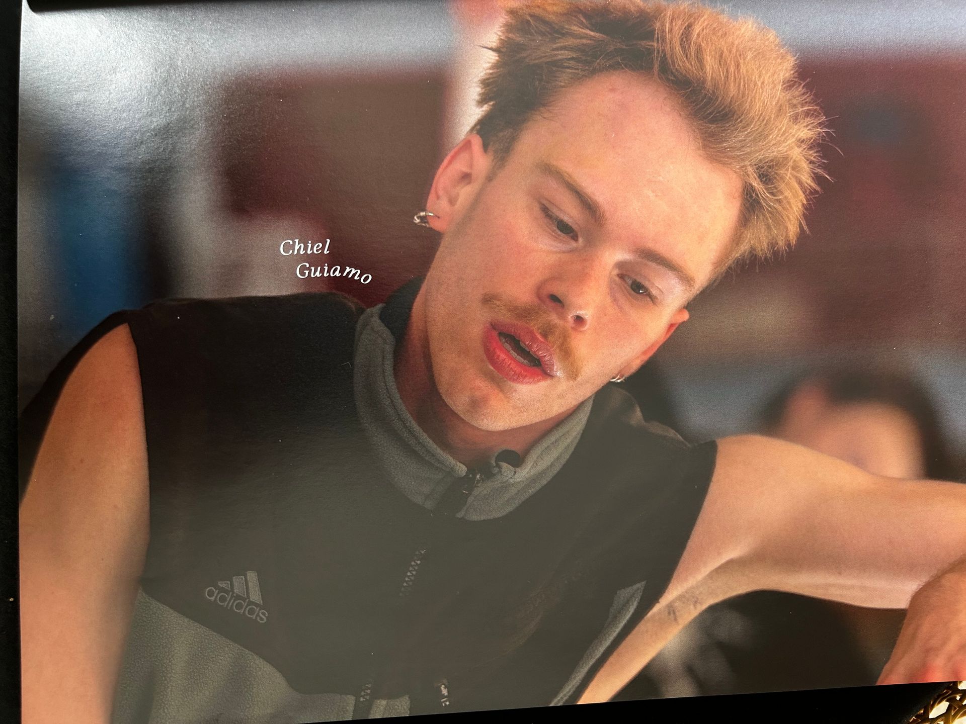 Een man met roodachtig haar en een snor, gekleed in een donker vest, kijkt naar beneden met open mond.