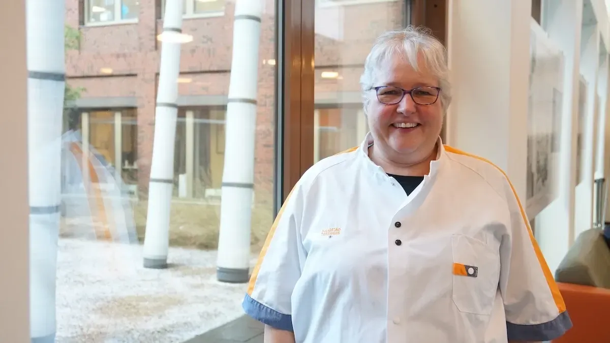 Een vrouw in een wit uniform staat glimlachend bij een raam met decoratieve bamboe. Oranje en blauwe accenten zijn zichtbaar.