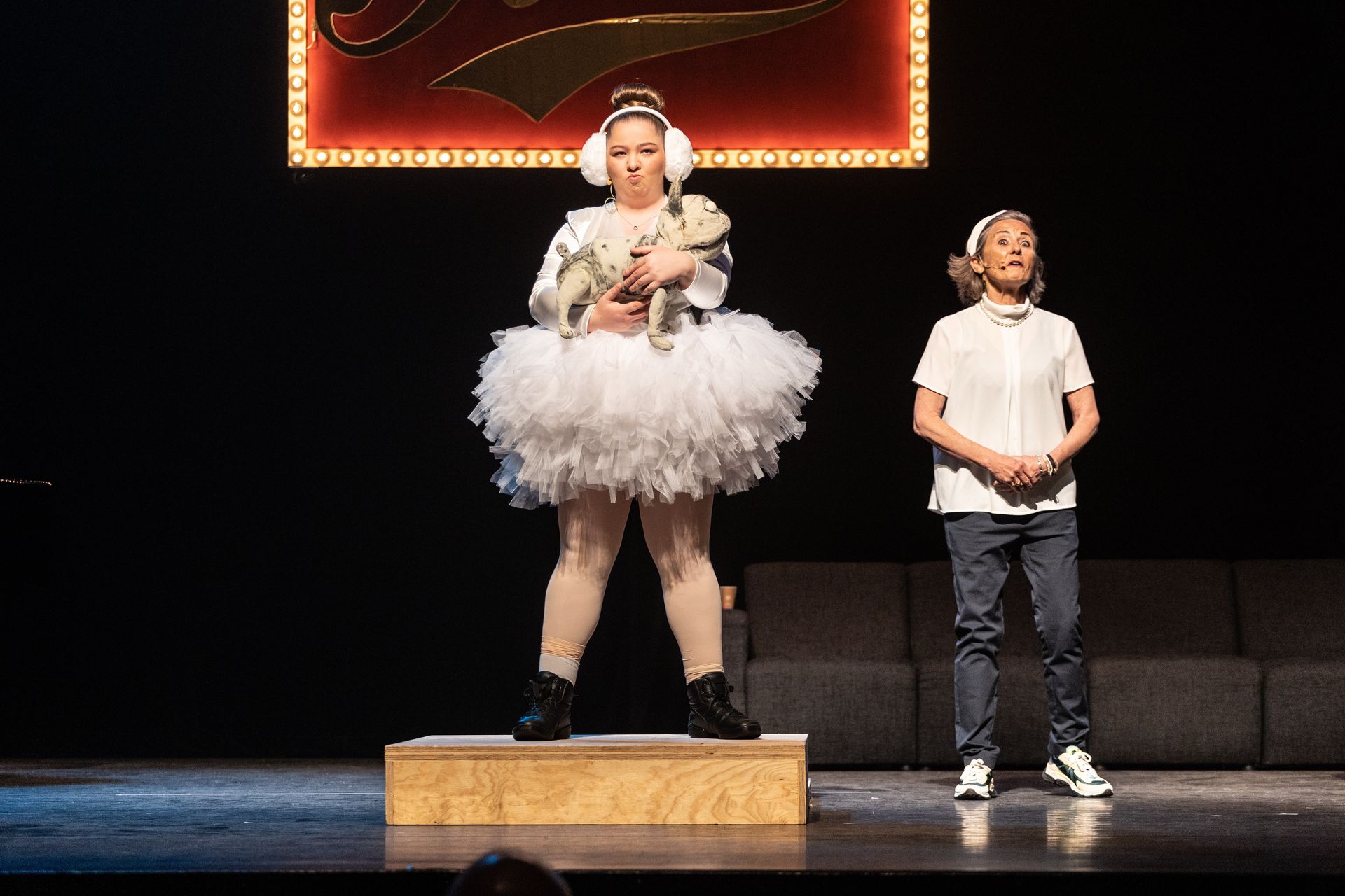 Twee mensen op het podium: een in een tutu met een pop in haar handen; een ander in een wit shirt en spijkerbroek.
