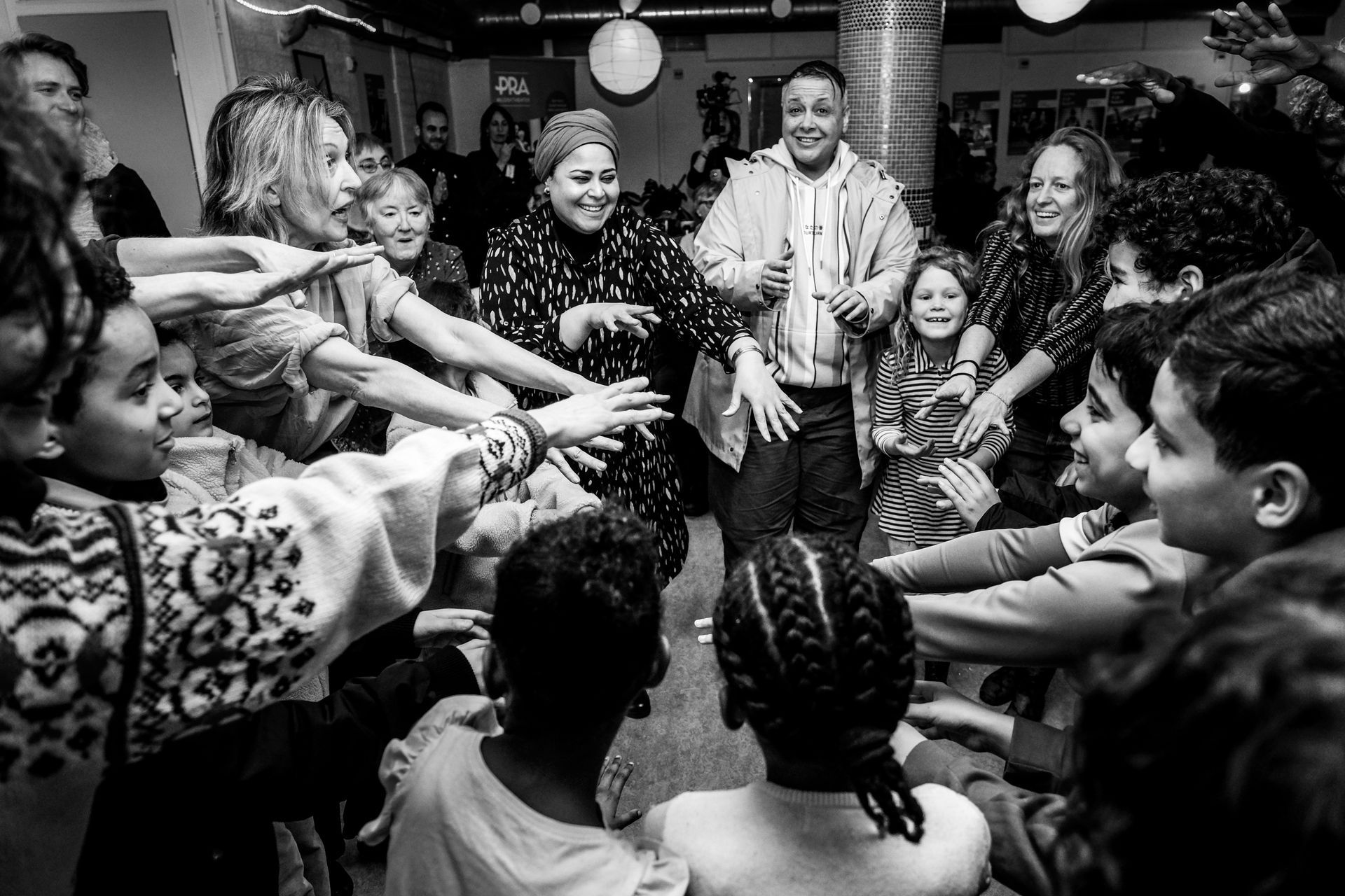 Mensen dansen in een kring, met de handen in elkaar. Binnen, met lachende volwassenen en kinderen. Zwart-wit.