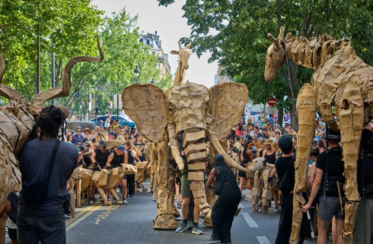 Straatparade met gigantische insectachtige poppen en een menigte toeschouwers op een met bomen omzoomde weg.