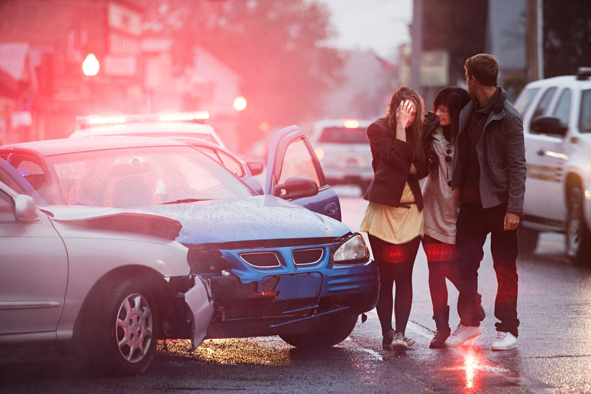 A car accident scene with a damaged vehicle, three people nearby, and flashing police lights