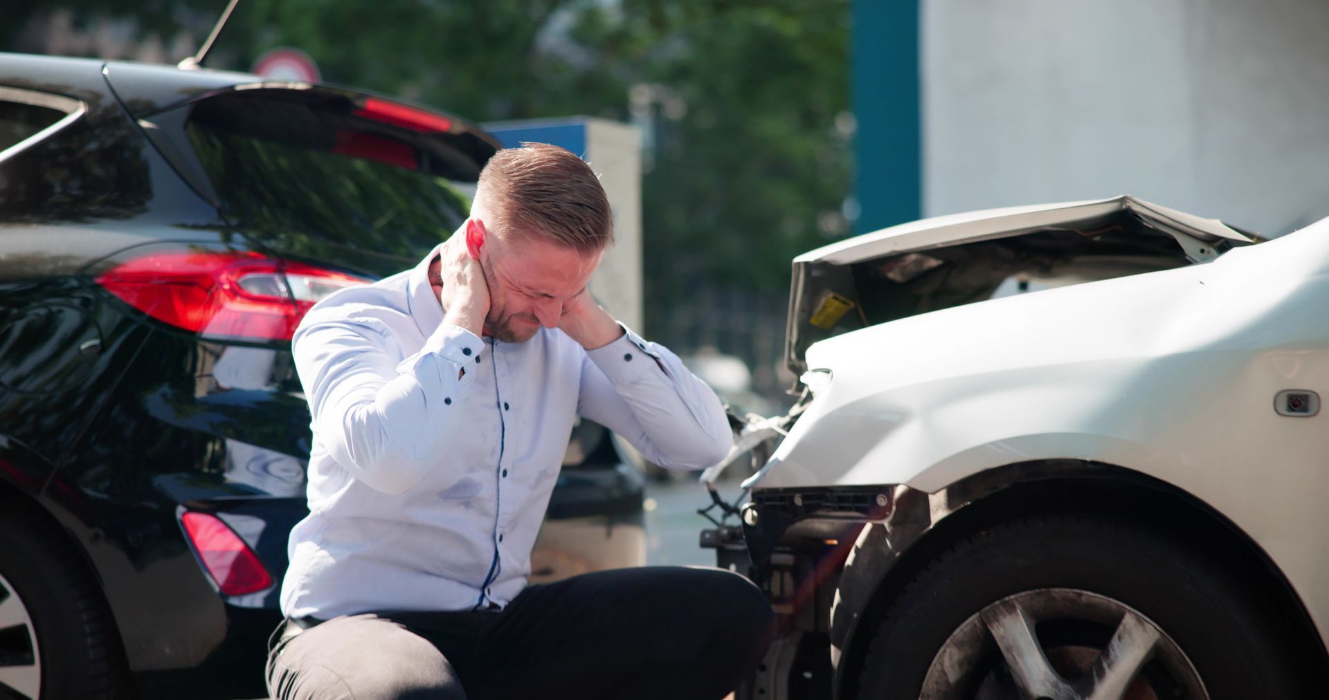 A man kneels by a car crash, holding his head in his hands, with damaged vehicles visible