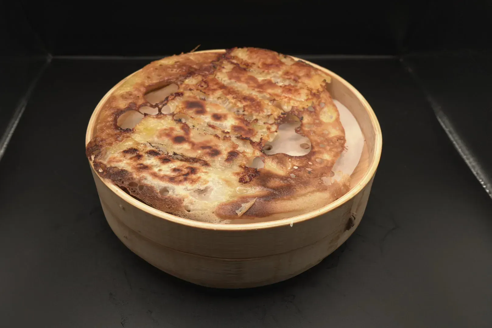 Round cake in a wooden bowl, topped with browned, mottled frosting on a dark background.