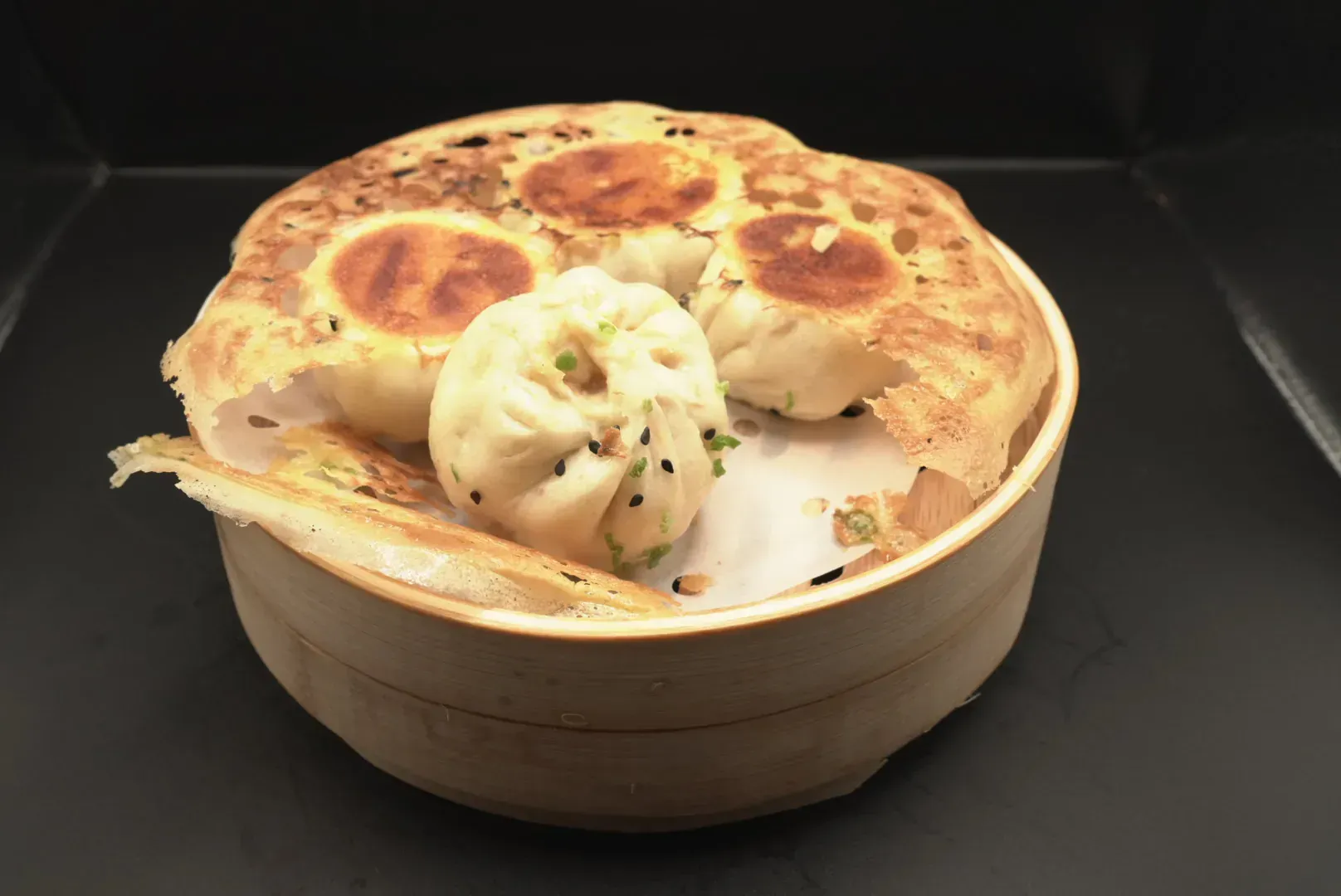 Round wooden basket with two golden-brown bao buns and one split dumpling on a dark background