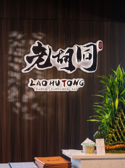 Restaurant sign on a dark wood wall reading “LAO HUI TONG,” with a plant and table in front