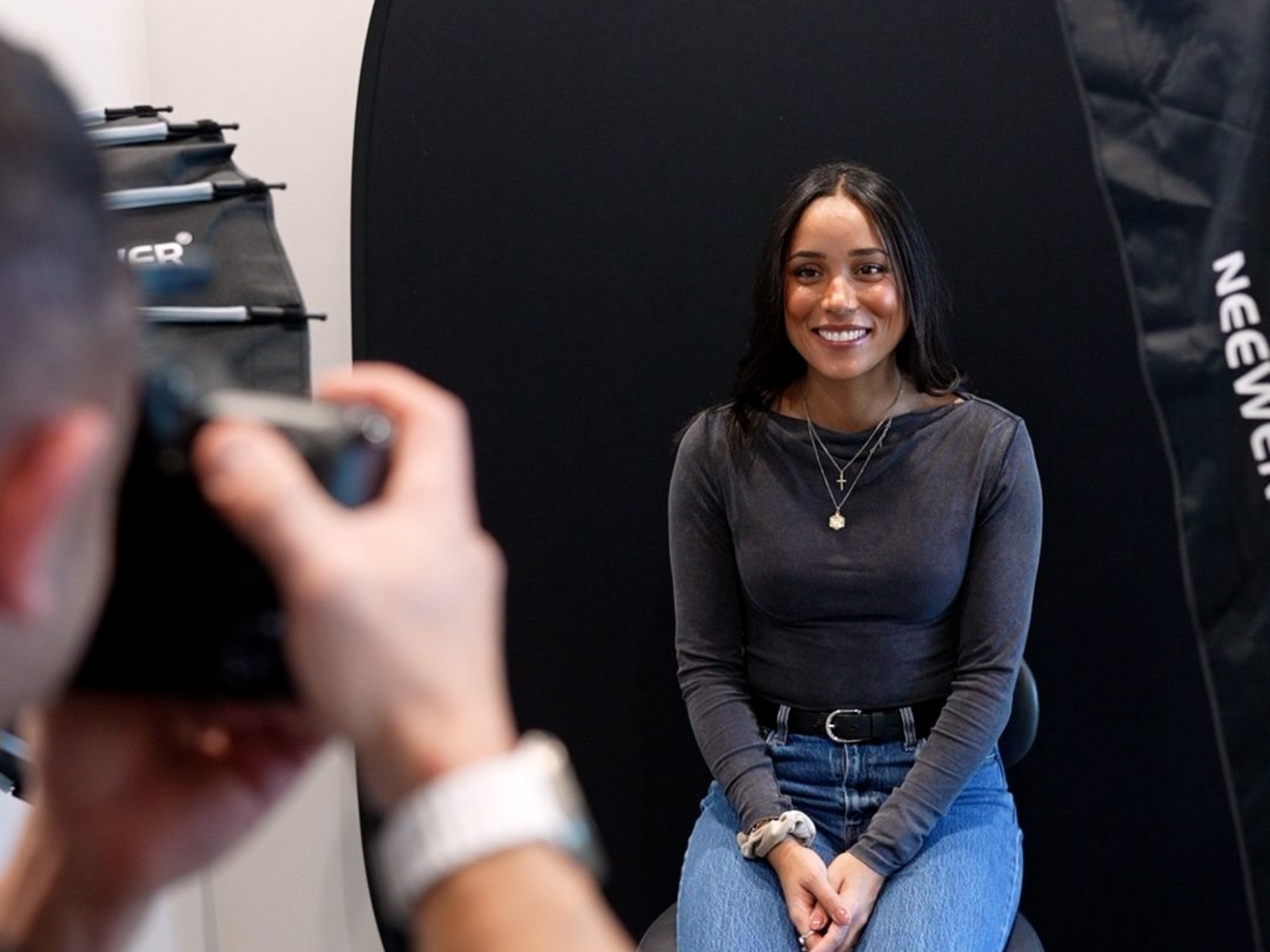 Person smiling in a studio portrait setup, seated against a black backdrop while being photographed.