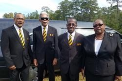 Four men in black suits with yellow striped ties standing outdoors by a lakeside.