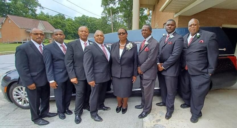 Eight people in business suits pose together outside a building, standing beside a parked car.