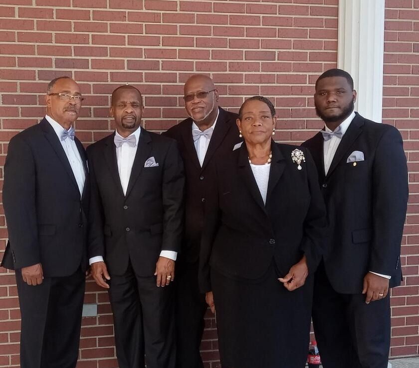 Five people in formal black suits posing in front of a brick wall