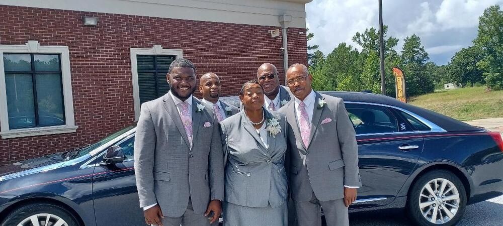 Five people in gray suits stand in front of a black sedan outside a brick building.