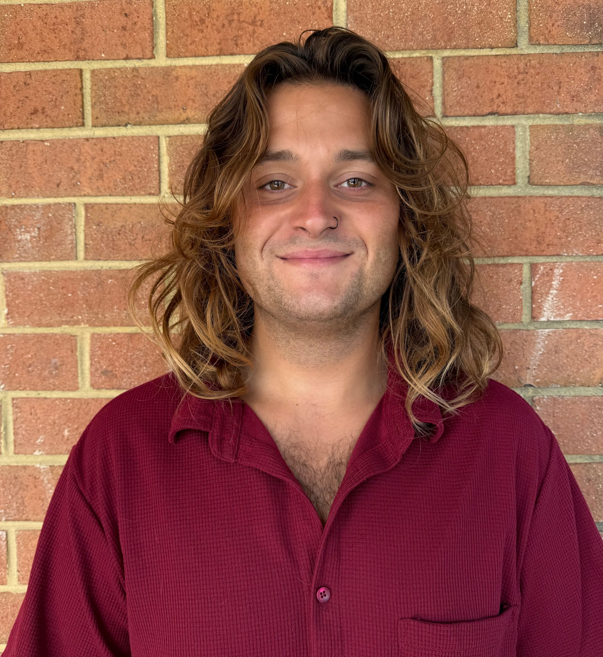 Man with long brown hair and nose ring, smiling in front of a white-sided building. He wears a tan striped shirt.