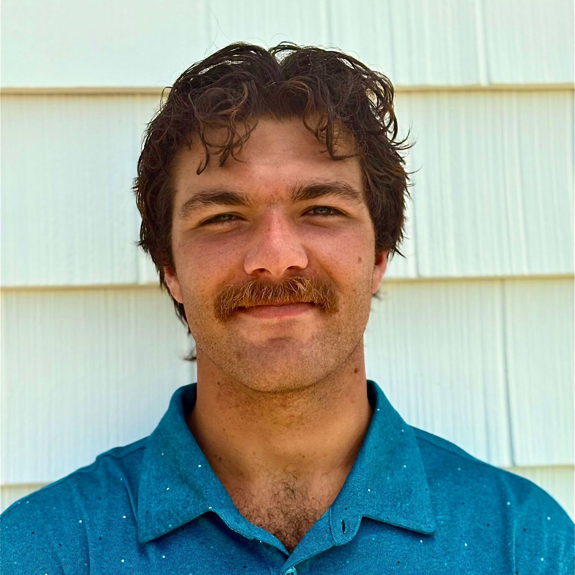 Man with brown hair and a mustache smiling, wearing a blue shirt, standing in front of a white shingled wall.