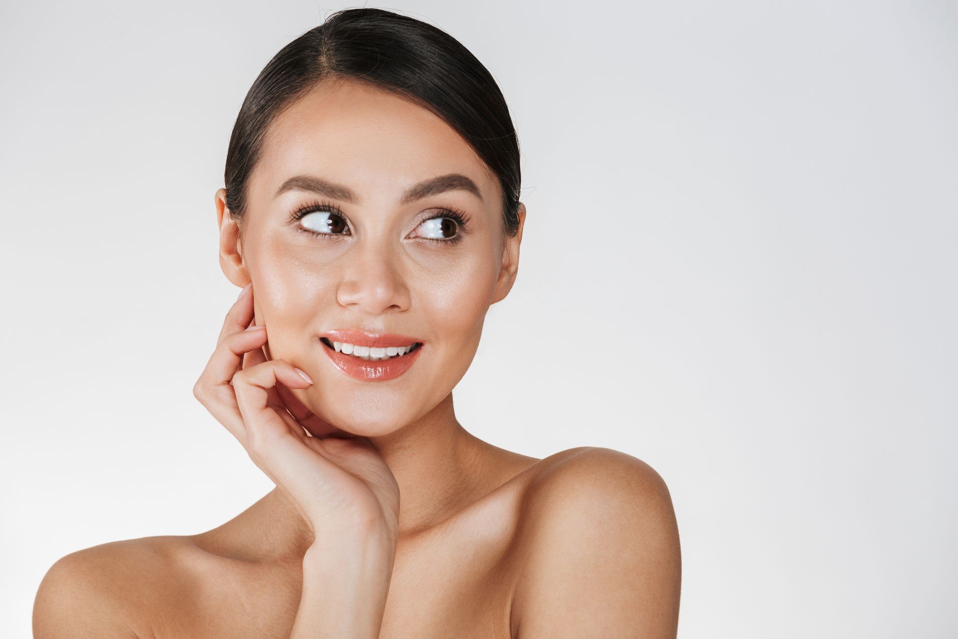 Woman with smooth skin smiles, gazing to the side, hand touching her cheek, against a white background.