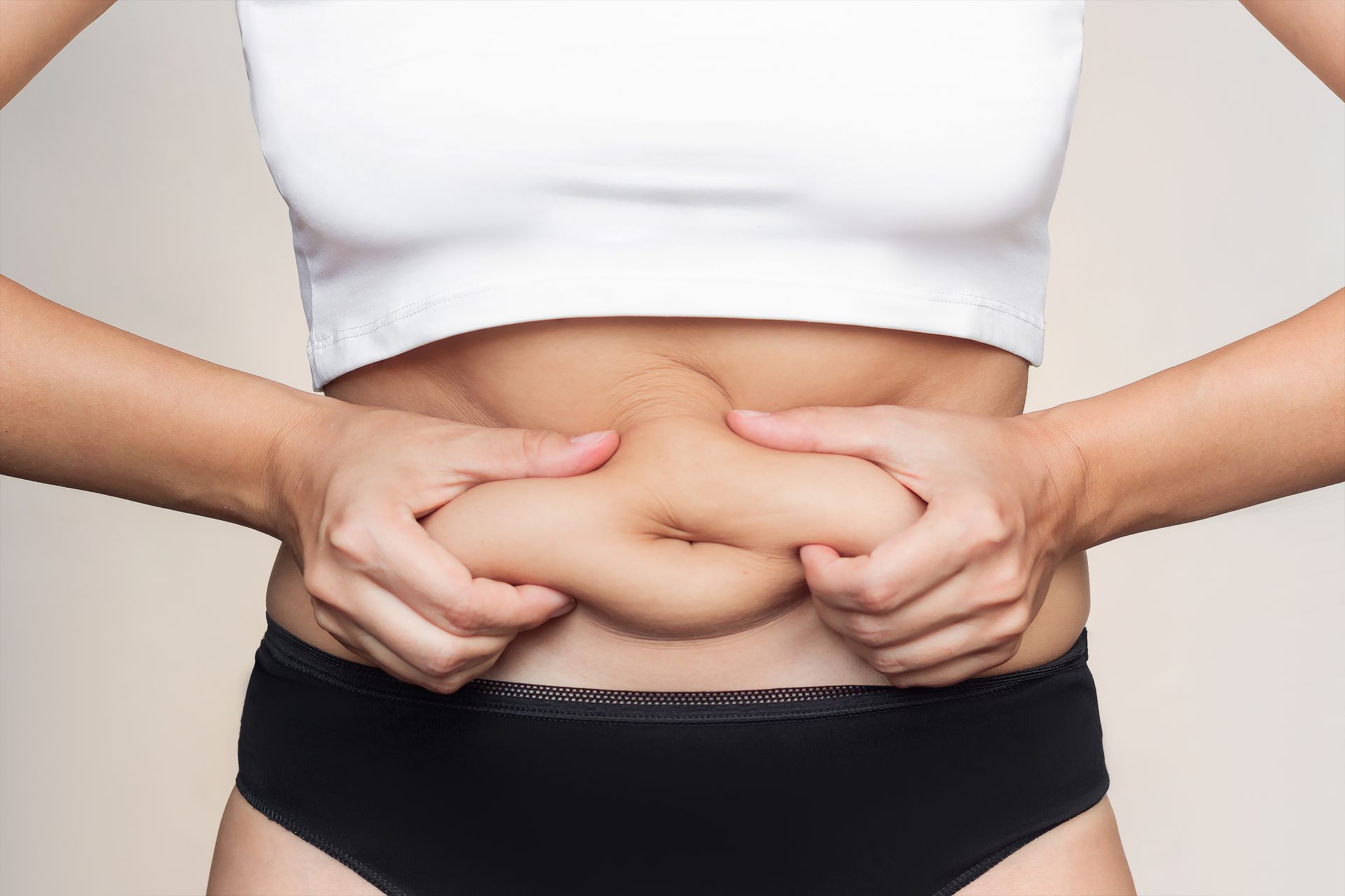 Woman holding and pinching her abdominal fat, wearing a white top and black underwear.