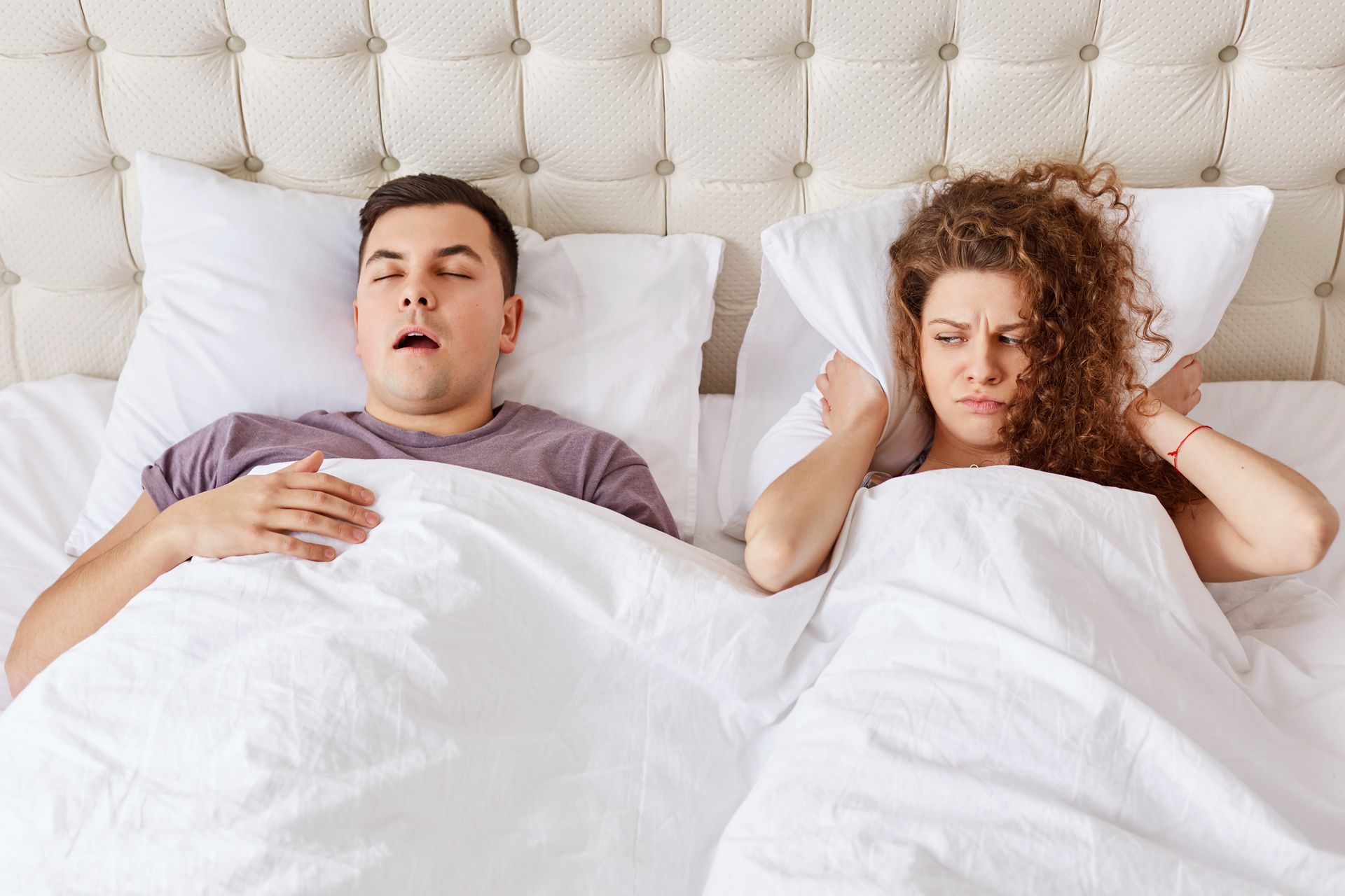 Man snoring in bed; woman covers ears with pillow, looking annoyed.