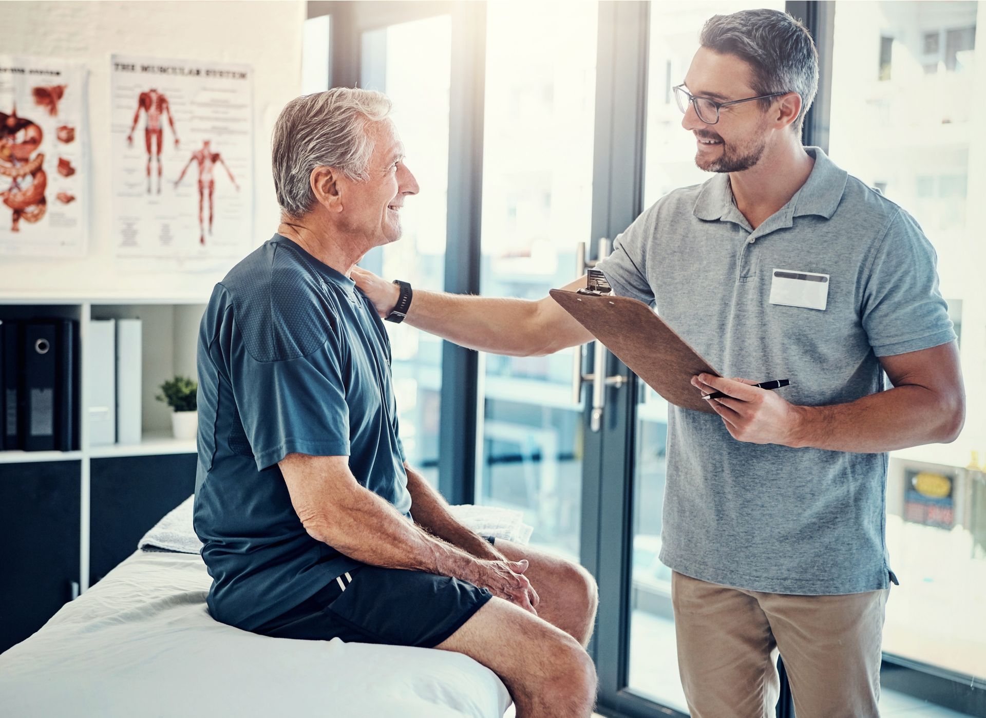 A physical therapist comforts a senior man, checking his chart. Inside a clinic, bright.
